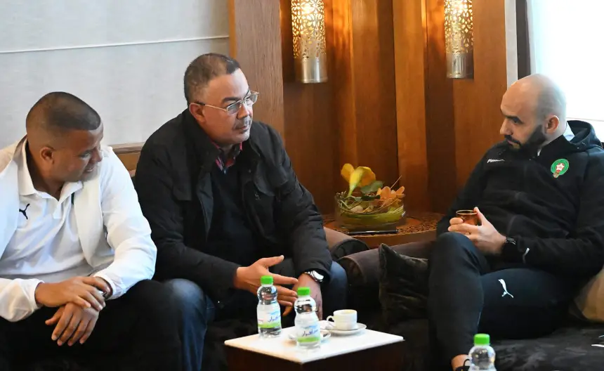 Trois hommes en tenue décontractée engagés dans une conversation, assis dans un cadre moderne avec des boissons sur une table, illustrant l'esprit d'équipe dans le football marocain.