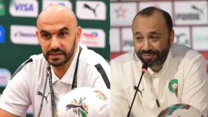 Twee voetbaltrainers in witte Puma-shirts, waarvan er één spreekt op een persconferentie, die het Marokkaanse voetbal vertegenwoordigen met het logo van het nationale team.