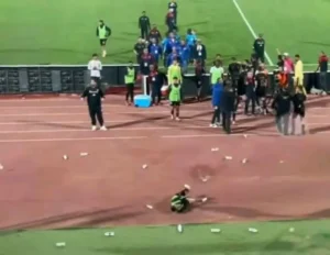 Fans celebrate on the sidelines as a Moroccan football player sits on the pitch. Bottles litter the ground, capturing the match's intensity.