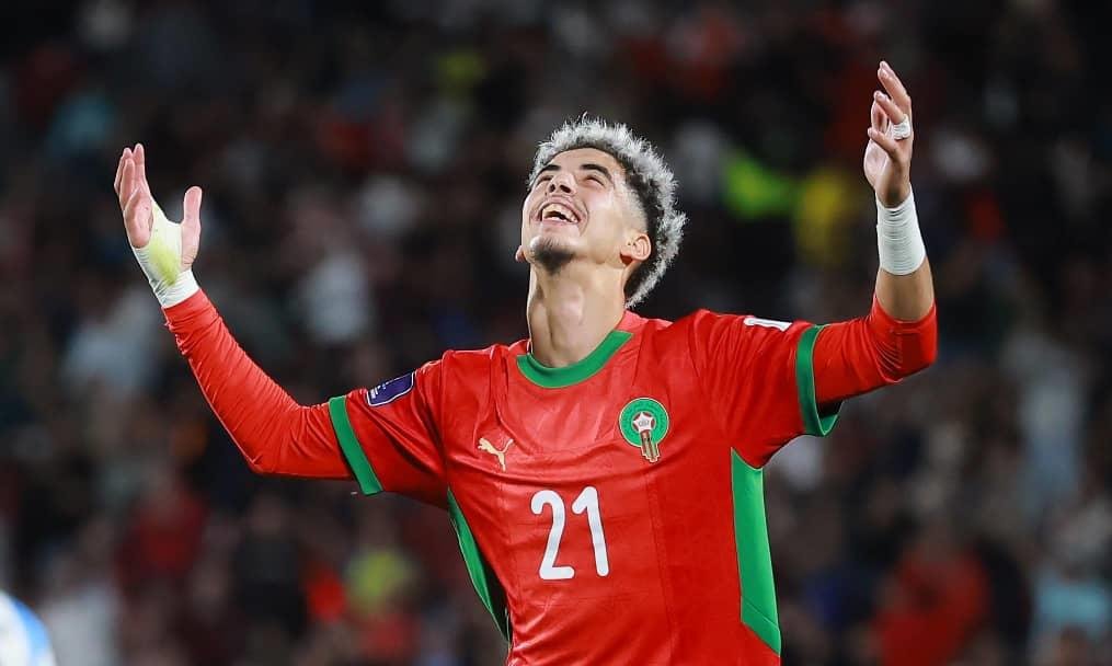 Moroccan player celebrating joyfully in a vibrant red kit with green trim, arms raised during an international match.