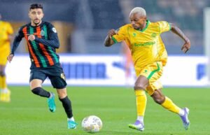 Football players in action: one in a yellow kit dribbling the ball, while the other in a black and green striped kit pursues him.