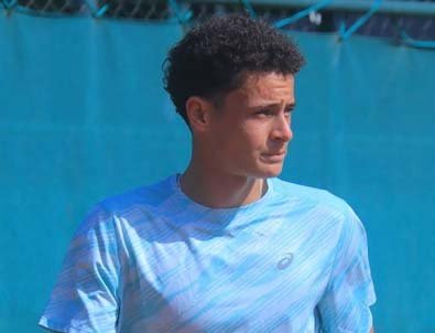 Young male tennis player with curly hair, wearing a blue striped shirt, stands on a court with a turquoise backdrop.