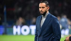 Moroccan footballer Mehdi Benatia in a navy suit, looking pensive at a match, with blurred teammates and stadium setting behind him.