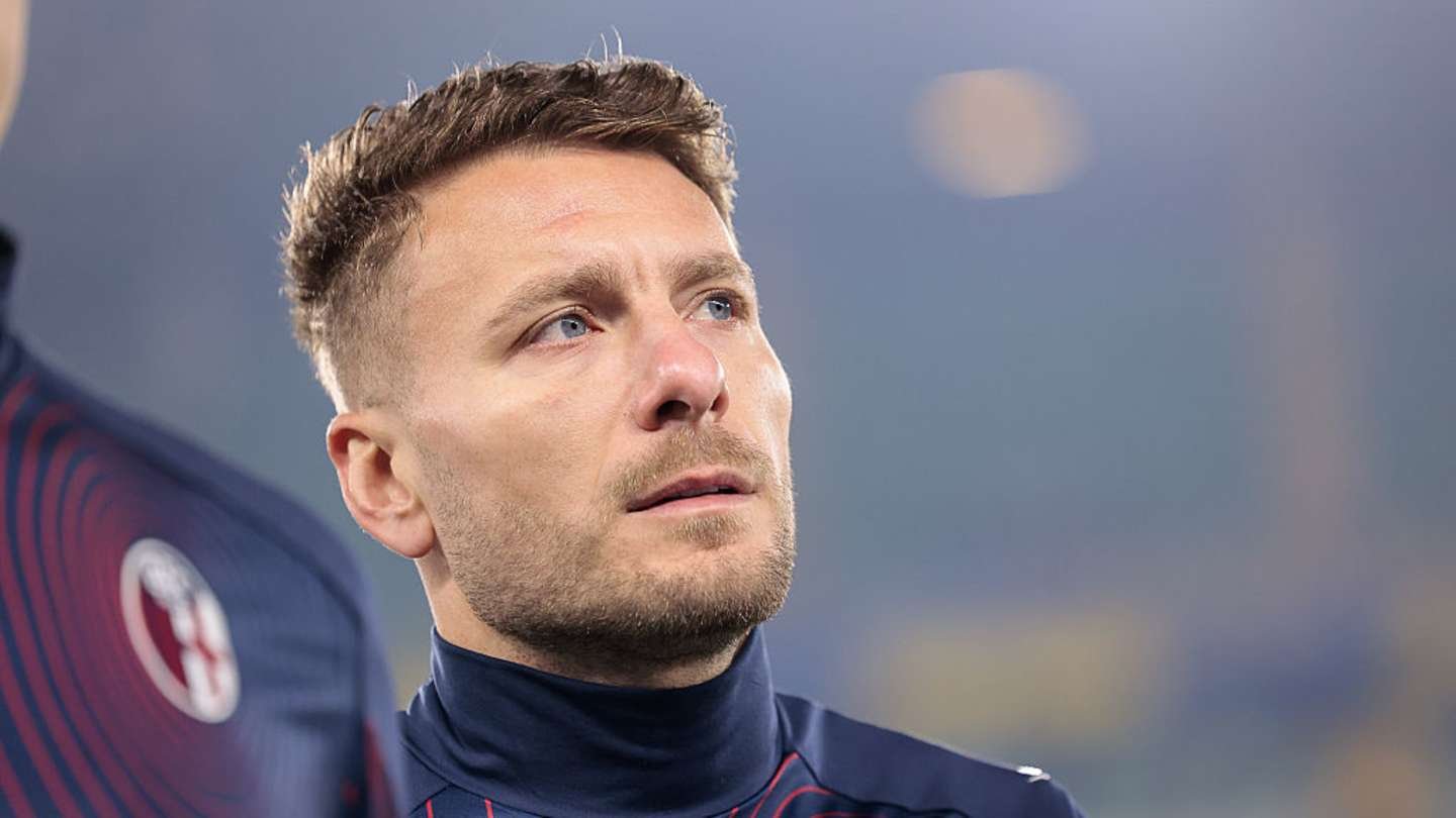 Crotone player gazes thoughtfully, wearing a dark training kit with club logo. Soft stadium lights create a moody atmosphere.