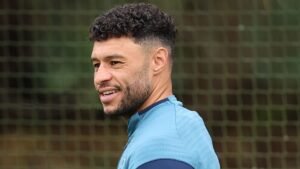Alex Oxlade-Chamberlain souriant pendant l'entraînement, portant un maillot bleu clair, avec un filet de football en arrière-plan.