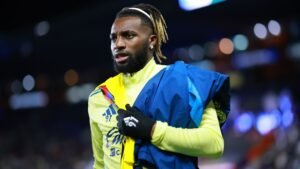 Allan Saint-Maximin portant le maillot jaune et bleu du Club América, tenant une veste, affichant une expression sérieuse pendant l'échauffement d'un match.