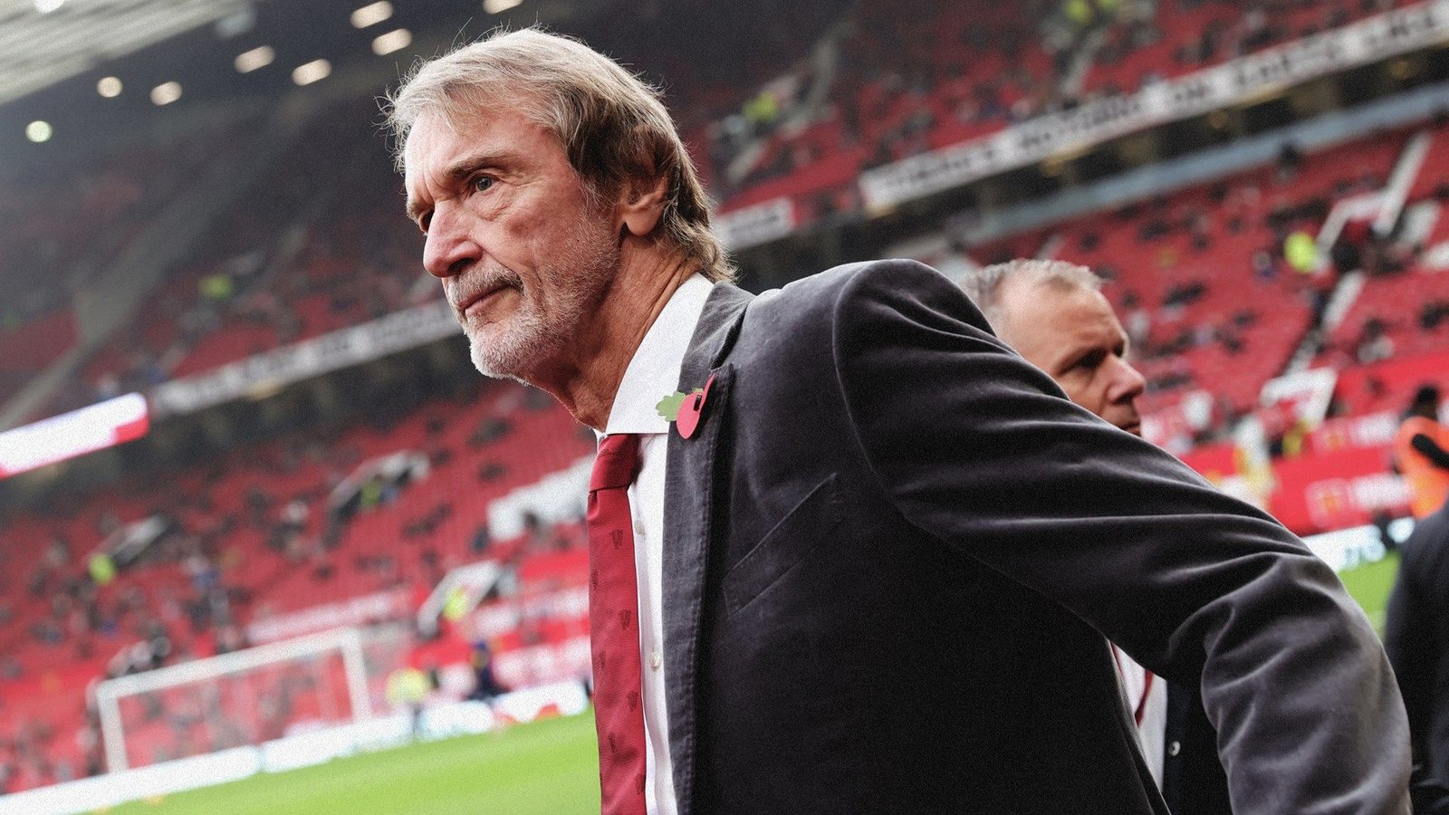 Zakenman Jim Ratcliffe in Old Trafford, gekleed in een donker pak met een klaproos, wandelend langs lege rode tribunes tijdens een wedstrijd.