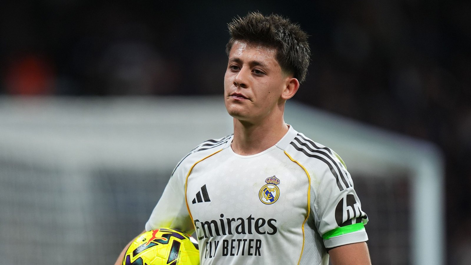 Arda Güler in het grijze tenue van Real Madrid, houdt een wedstrijdbal vast met een serieuze uitdrukking tijdens een gespannen moment op het veld.