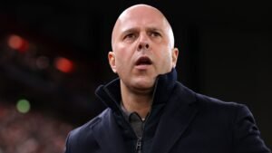 Arne Slot apparaît déterminé et concentré lors d'un match, portant un manteau sombre, avec un fond de stade flou.