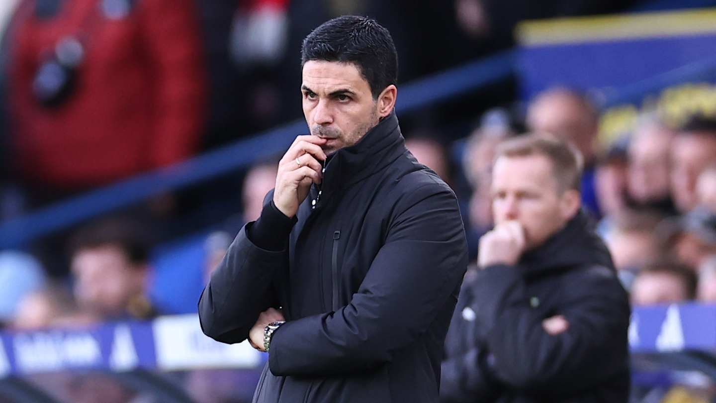 Mikel Arteta in a black jacket, pensive expression, observing play during a match, with coaching staff in the background.
