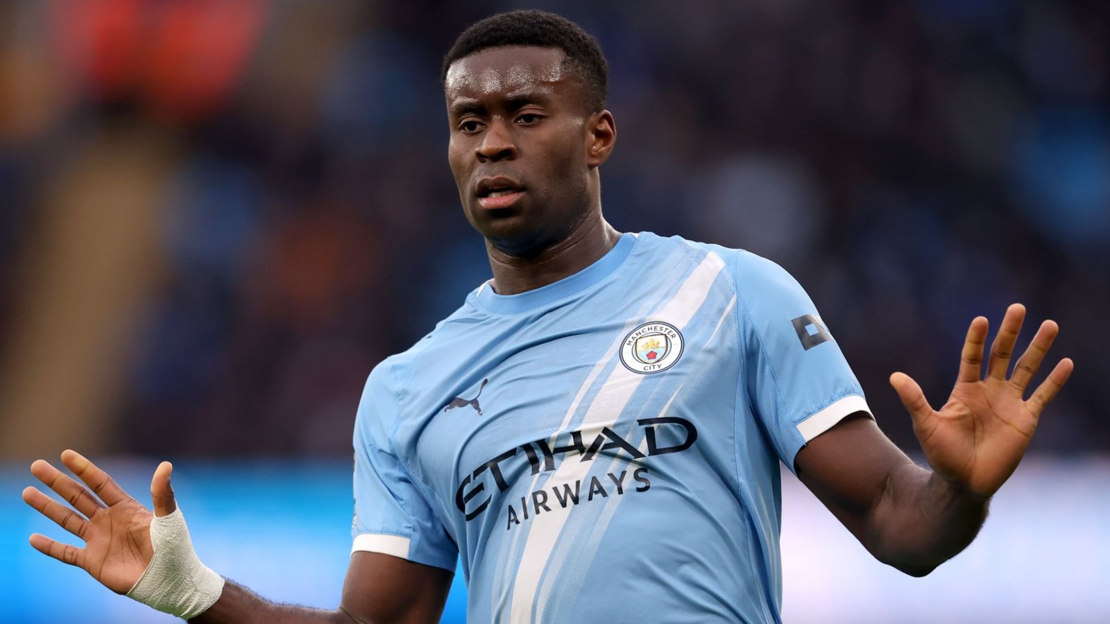 Nathan Aké dans le maillot bleu clair de Manchester City, gesticulant pendant un match, démontrant sa concentration et son leadership sur le terrain.