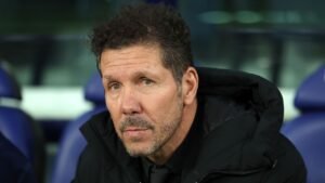 Diego Simeone, in a black jacket, pensive expression, seated on the bench during a match, reflecting on his future at Atletico Madrid.