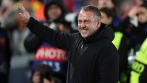 Barcelona's new striker coach smiles and waves at the crowd during a match celebration, wearing a black jacket against a lively backdrop.