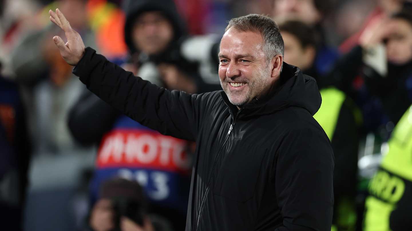 Barcelona's new striker coach smiles and waves at the crowd during a match celebration, wearing a black jacket against a lively backdrop.