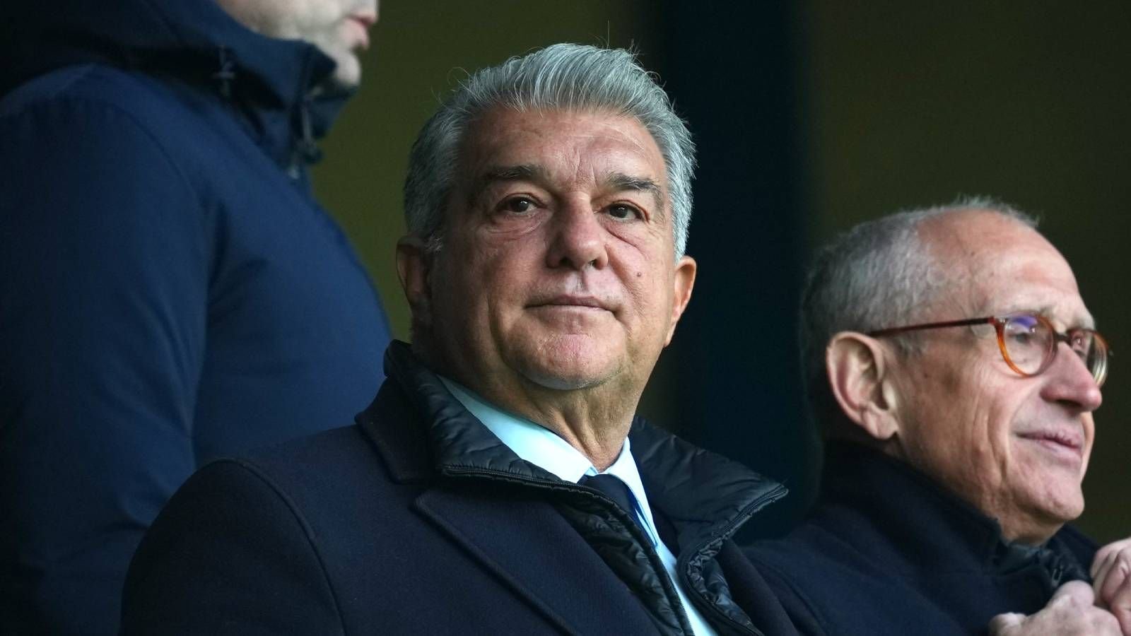 Joan Laporta, président du FC Barcelone, assis avec un collaborateur, tous deux en tenue formelle, regardant un match dans un stade.
