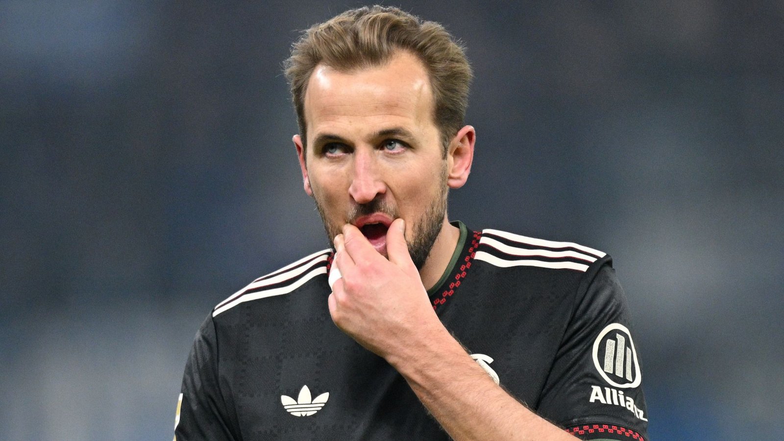 Harry Kane, portant la tenue noire du Bayern Munich, semble préoccupé en touchant sa bouche pendant un match, soulignant sa récente maladie.