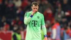Bayern Munich goalkeeper Manuel Neuer, in a green kit, shows disappointment during a match, with fans and players blurred in the background.