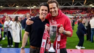 Ben Foster and Ryan Reynolds celebrate holding a trophy on the pitch at Wrexham, surrounded by fans in a vibrant stadium atmosphere.