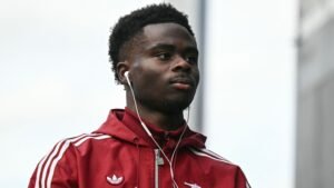 Bukayo Saka in een rood Arsenal-trainingspak, met oortjes in, lopend richting het stadion voorafgaand aan de wedstrijd tegen Leeds.