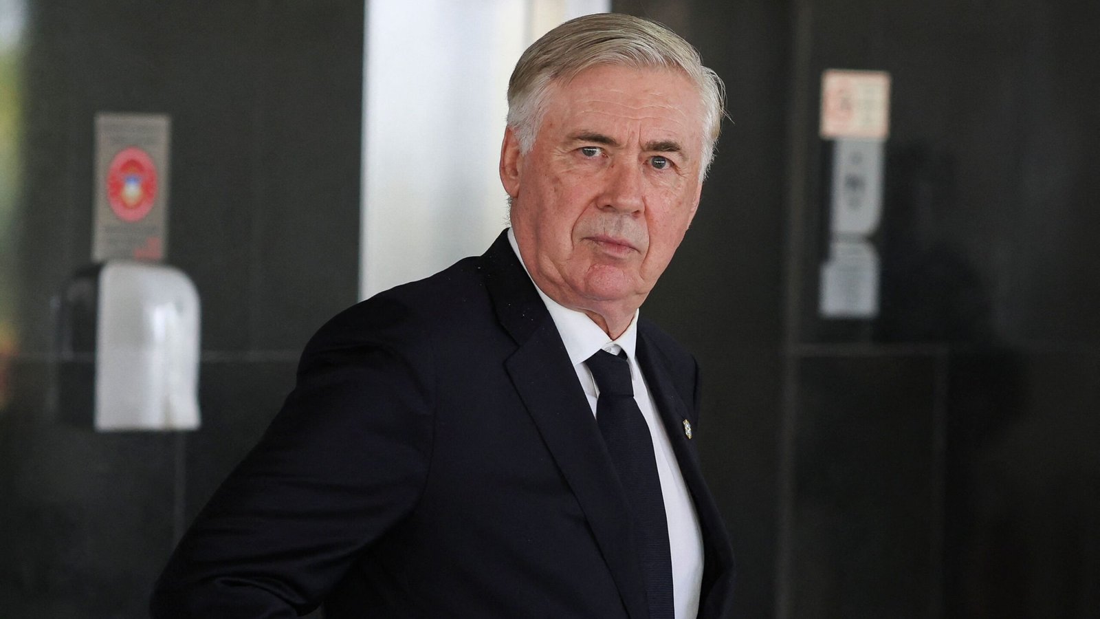 Carlo Ancelotti in formal attire, looking serious as he walks, with a modern building backdrop.