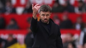 Michael Carrick gesticule en bord de touche lors d'un match à Old Trafford, portant un manteau sombre devant un fond de tribunes rouges.