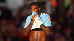 Un joueur de football aux cheveux tressés s'essuie le visage avec une serviette, portant un maillot bleu clair, et montre des émotions pendant un match.