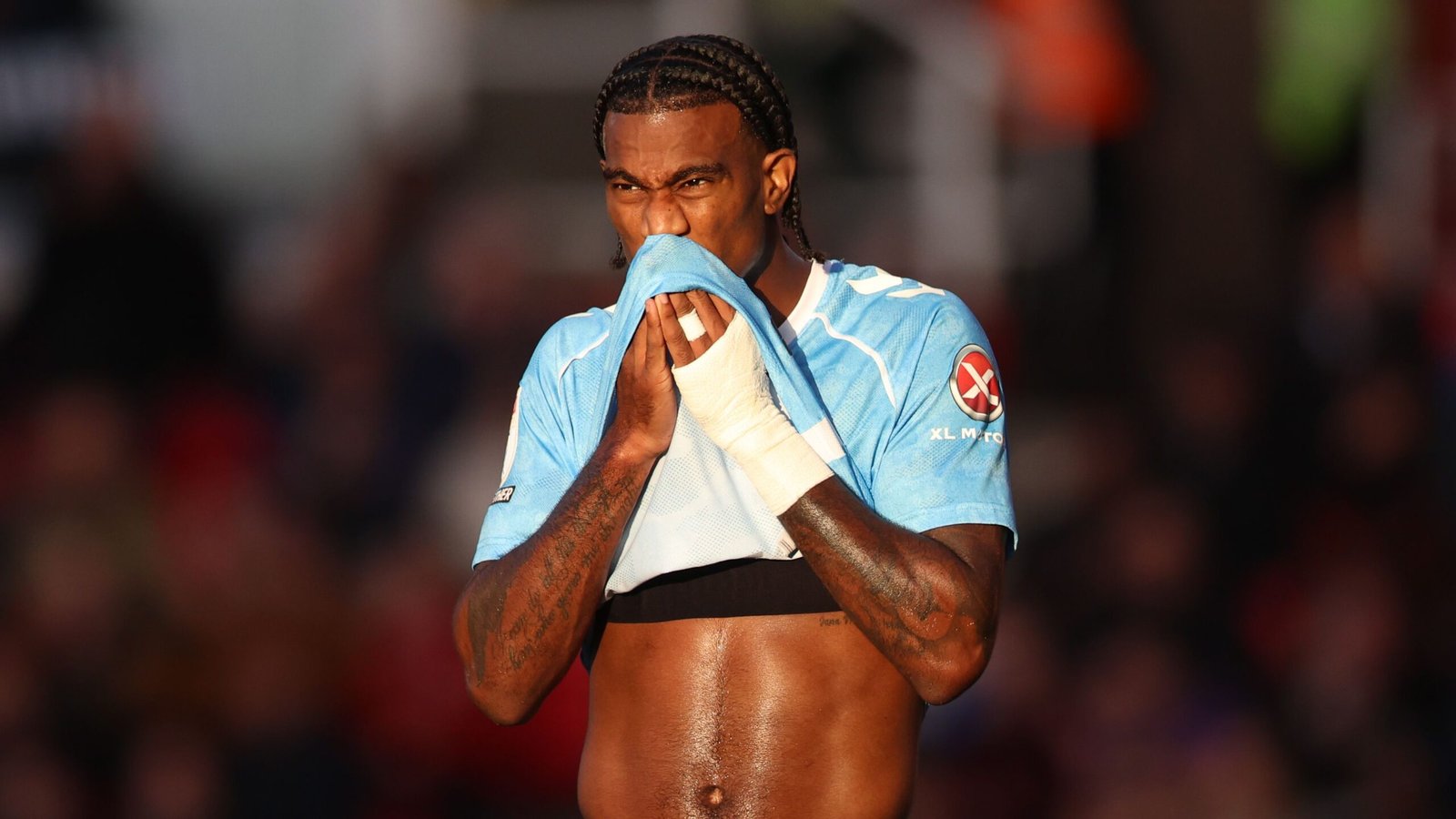 Un joueur de football aux cheveux tressés s'essuie le visage avec une serviette, portant un maillot bleu clair, et montre des émotions pendant un match.