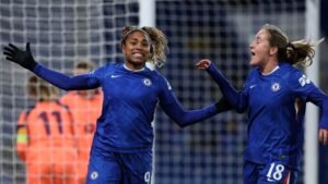 Catarina Macario viert een doelpunt met een teamgenoot, beiden in het blauwe tenue van Chelsea, met het doelnet en fans op de achtergrond.