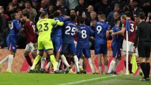 Les joueurs de Chelsea et de West Ham engagés dans une violente altercation pendant un match, entourés de supporters dans un stade animé.