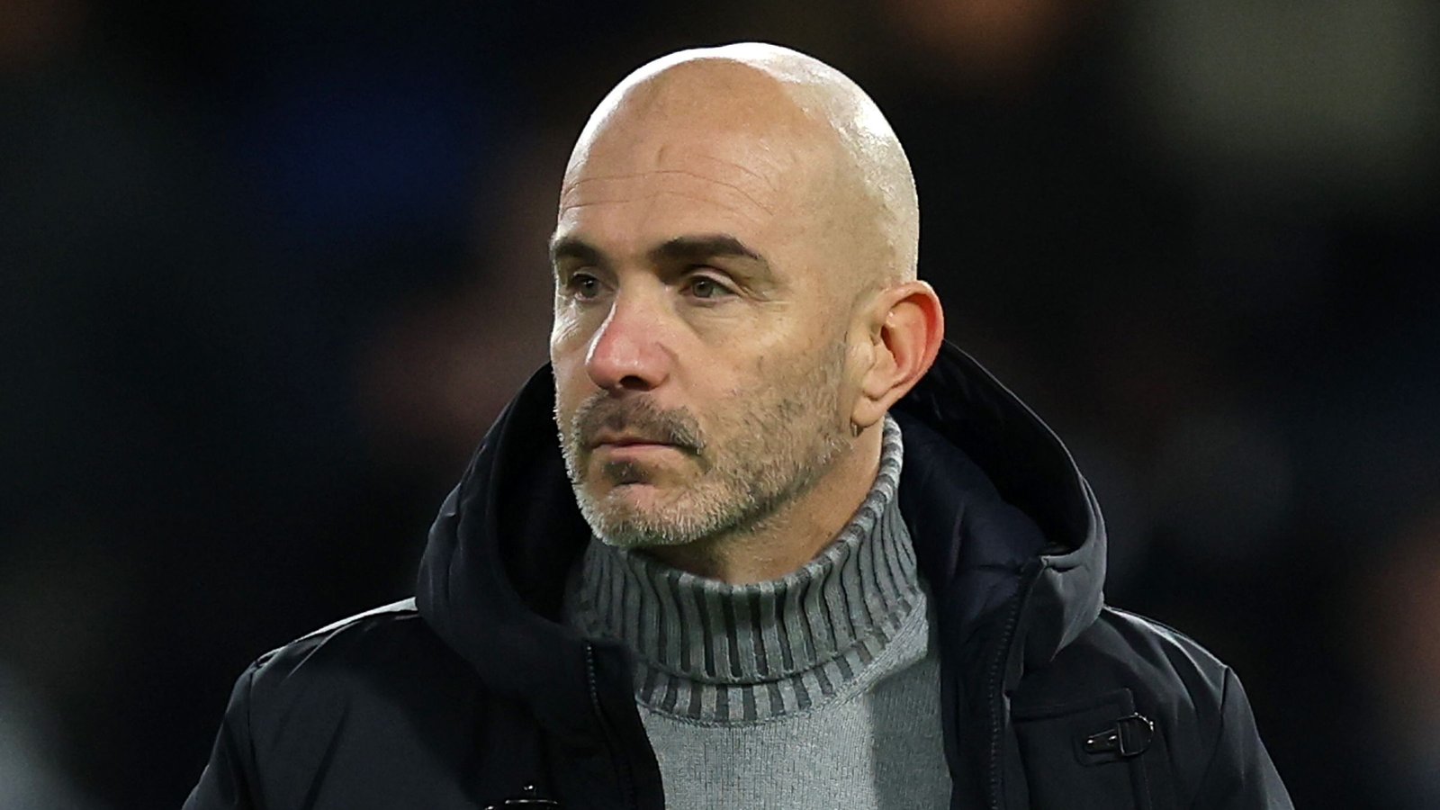 L'entraîneur de Chelsea, l'air pensif, veste sombre et col roulé gris, devant un fond de stade flouté.