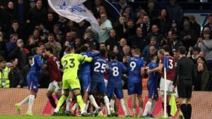 Chelsea-spelers in blauwe tenues vieren een doelpunt te midden van een levendige menigte, terwijl op de achtergrond een spandoek van fans wappert tijdens een wedstrijd.