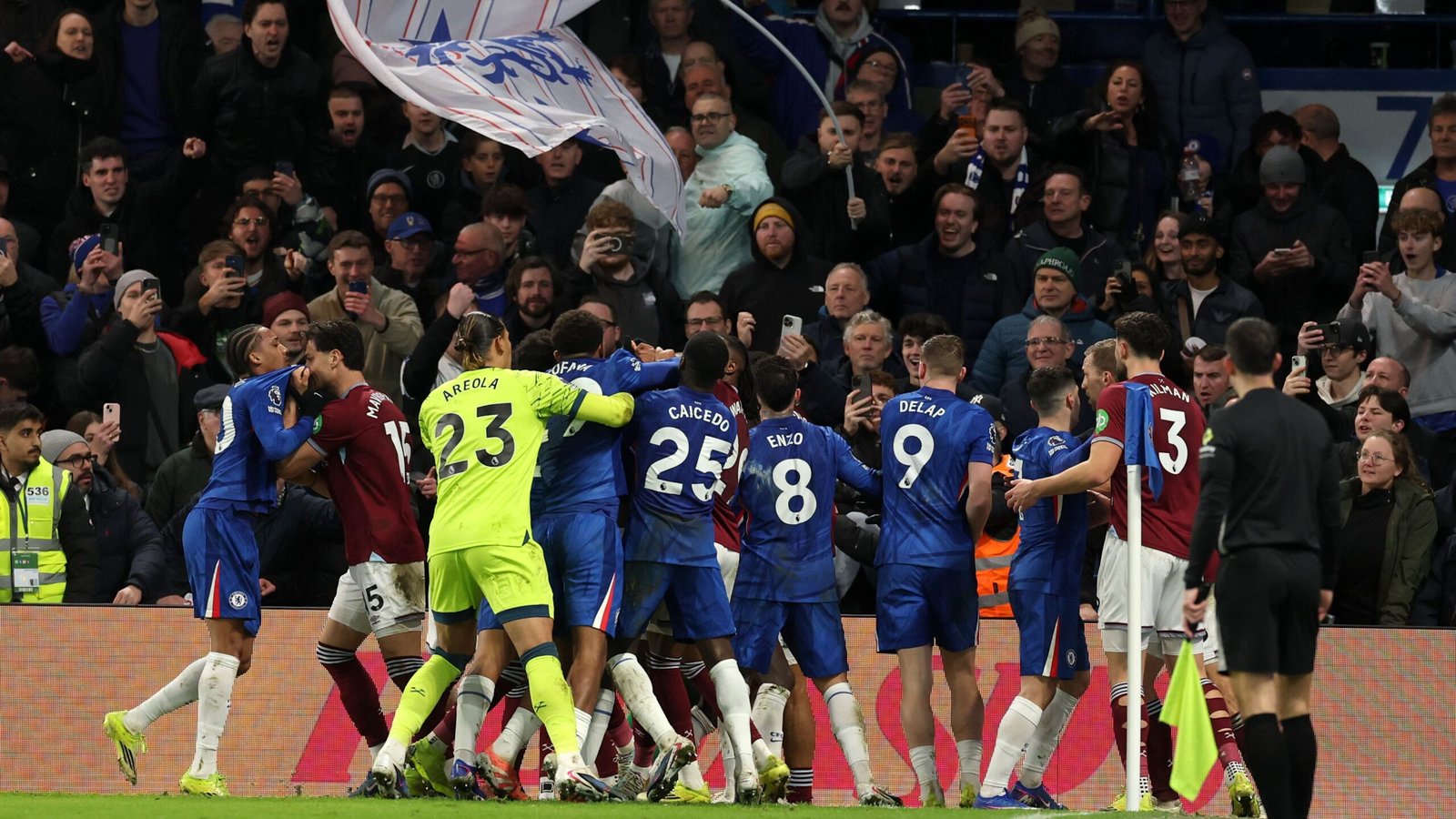 Chelsea-spelers in blauwe tenues vieren een doelpunt te midden van een levendige menigte, terwijl op de achtergrond een spandoek van fans wappert tijdens een wedstrijd.