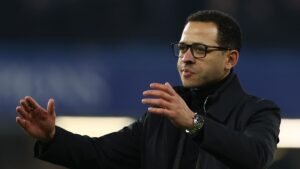 L'entraîneur de football marocain gesticule pendant un match, portant des lunettes et un manteau noir, concentré sur la stratégie de l'équipe.