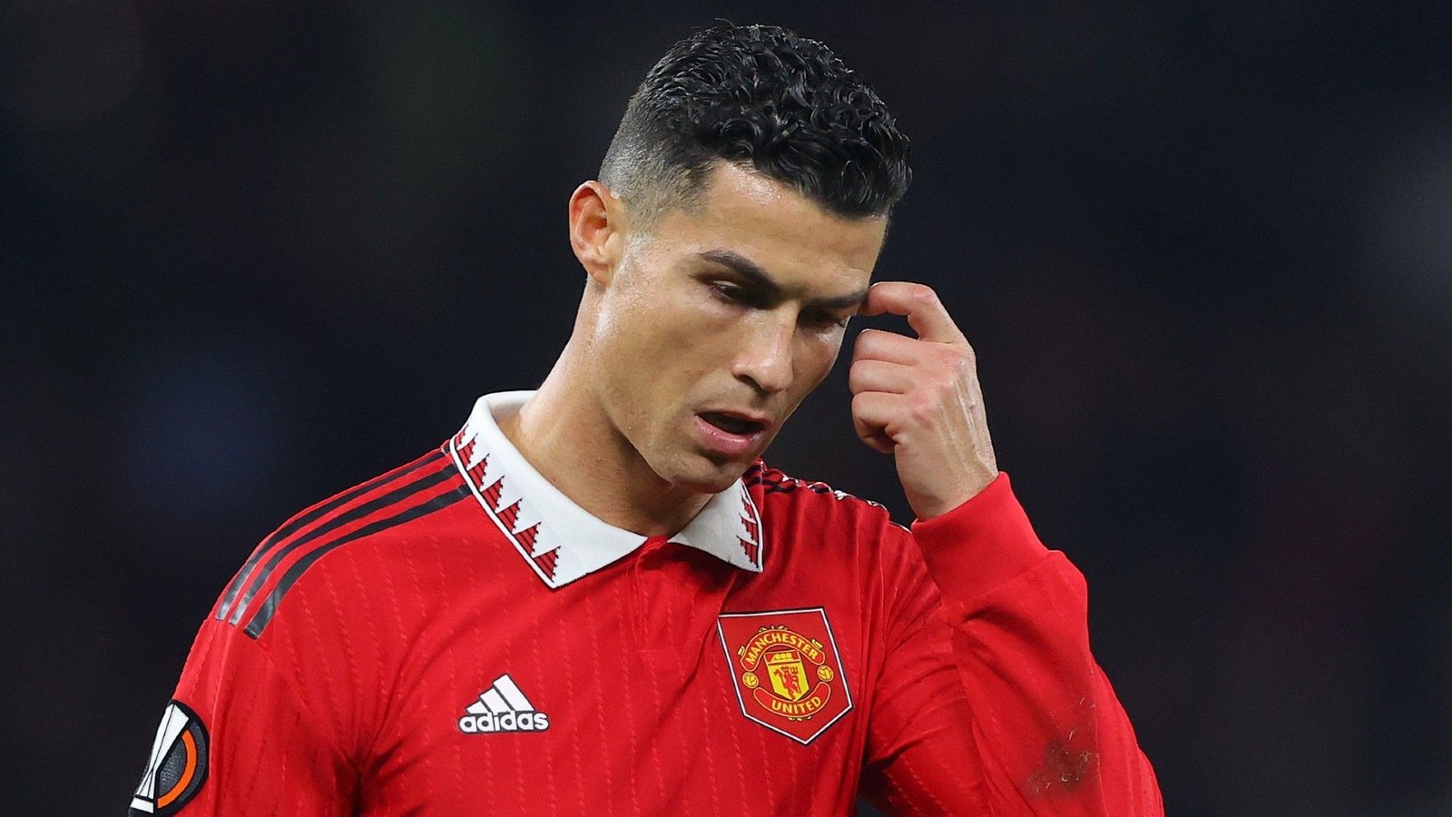 Cristiano Ronaldo in een rood Manchester United-tenue met een bedachtzame uitdrukking tijdens een wedstrijd, waarbij de clubbranding en focus worden getoond.