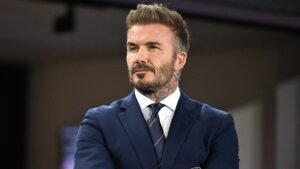 David Beckham, en costume bleu marine, arborant une barbe soignée et des tatouages, observe pensivement, incarnant son avenir potentiel en tant que propriétaire de club.