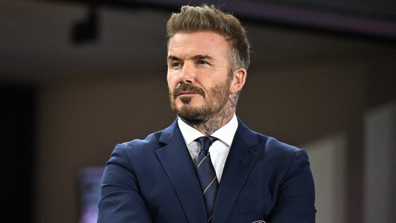 David Beckham, en costume bleu marine, arborant une barbe soignée et des tatouages, observe pensivement, incarnant son avenir potentiel en tant que propriétaire de club.