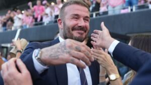 David Beckham souriant et célébrant avec des fans, vêtu d'un costume, dans une atmosphère de stade vibrante.