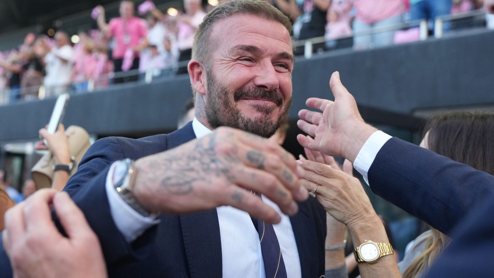 David Beckham souriant et célébrant avec des fans, vêtu d'un costume, dans une atmosphère de stade vibrante.