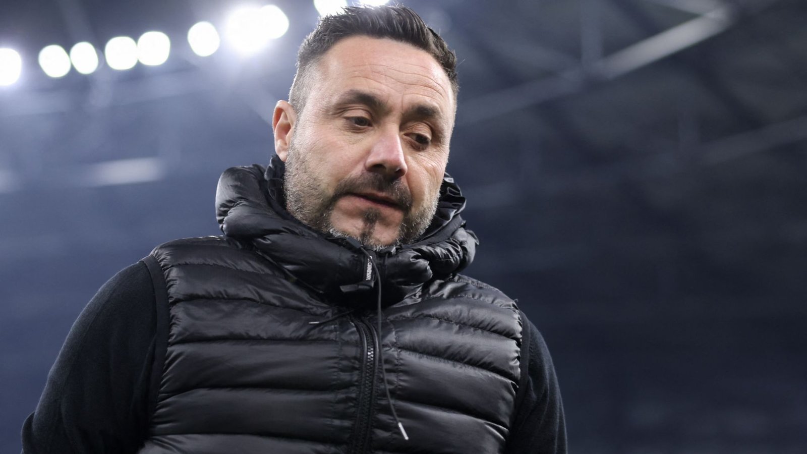 De Zerbi, vêtu d'une doudoune noire, semble contemplatif après la défaite de Marseille contre le PSG, sous les projecteurs du stade.