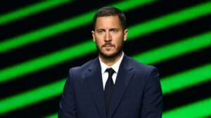 Eden Hazard, vêtu d'un costume bleu marine élégant, se tient devant un fond rayé vert, dégageant une assurance palpable lors d'une cérémonie de remise de prix footballistiques.