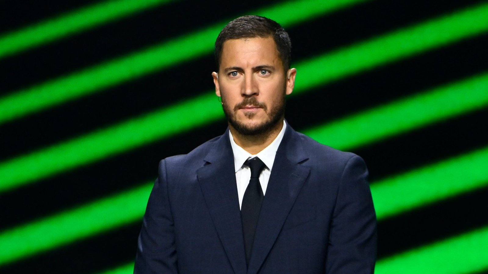 Eden Hazard, vêtu d'un costume bleu marine élégant, se tient devant un fond rayé vert, dégageant une assurance palpable lors d'une cérémonie de remise de prix footballistiques.
