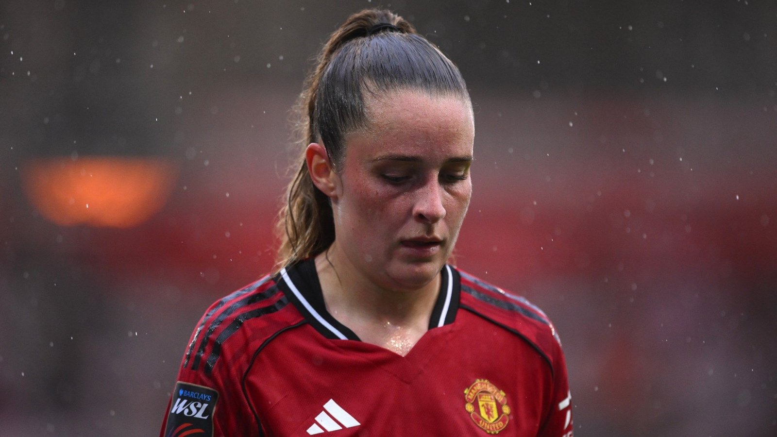 Ella Toone in een Manchester United-shirt, somber kijkend tijdens een regenachtige wedstrijd, wat haar teleurstelling over het missen van belangrijke wedstrijden benadrukt.