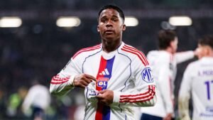 Lyon player showing his jersey, celebrating a moment on the pitch during a Ligue 1 match, with teammates in the background.