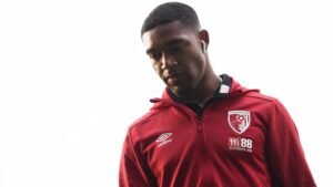 Jordon Ibe wearing a red Bournemouth tracksuit, looking down with earbuds, capturing a moment of contemplation before a match.