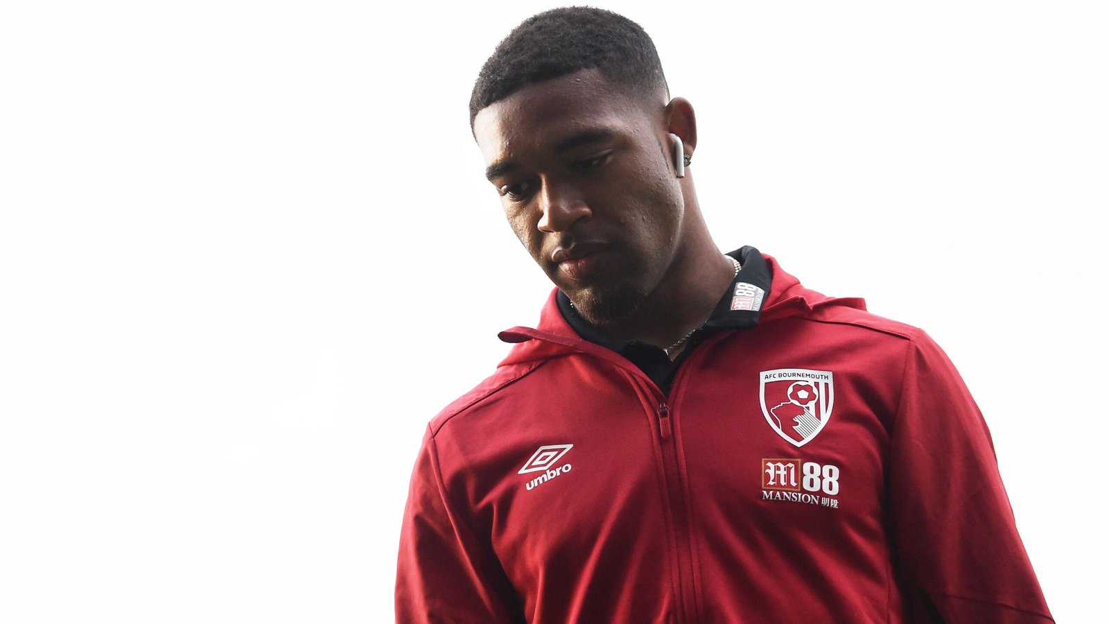 Jordon Ibe wearing a red Bournemouth tracksuit, looking down with earbuds, capturing a moment of contemplation before a match.