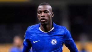 Chelsea player in a blue kit appears focused during a match, featuring the club crest prominently on his chest.