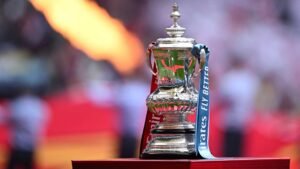 FA Cup trophy with a blue and red ribbon, perched on a podium against a blurred stadium background. Celebrating football heritage.