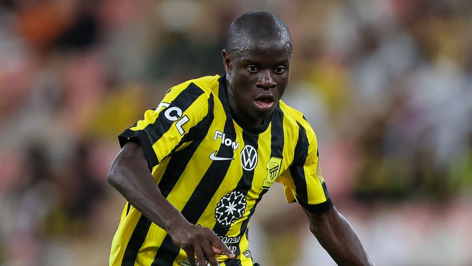 Le joueur d'Al Ittihad, N'Golo Kanté, portant un maillot rayé jaune et noir frappant, affichant une concentration intense pendant un match.