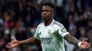 Vinícius Júnior célèbre un but pour le Real Madrid, portant le maillot blanc aux accents noirs du club, les bras grands ouverts en signe de triomphe.