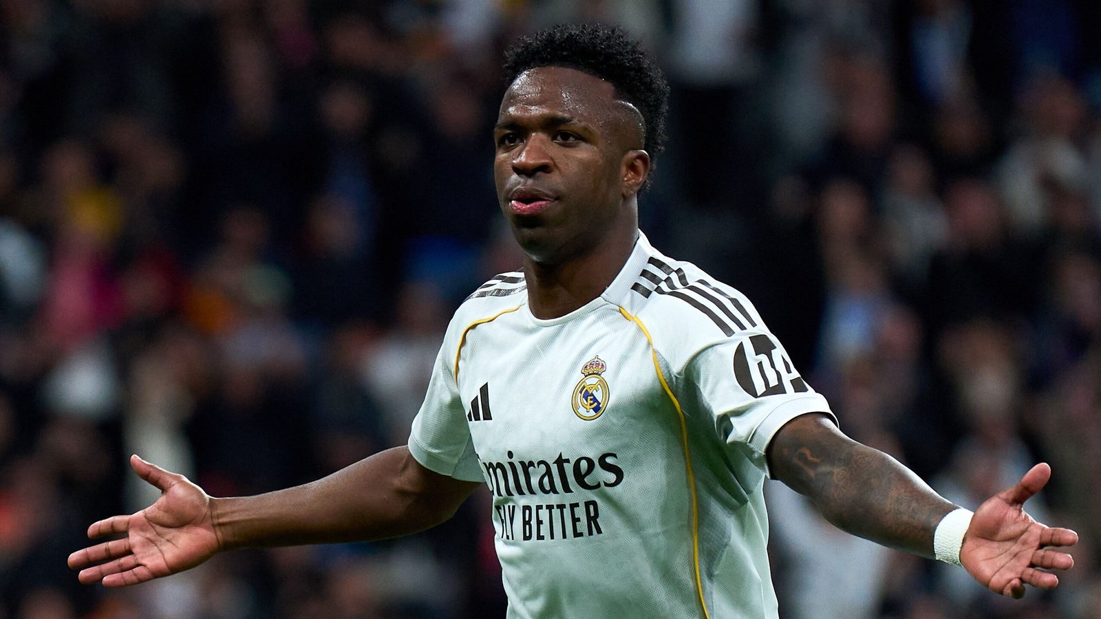 Vinícius Júnior célèbre un but pour le Real Madrid, portant le maillot blanc aux accents noirs du club, les bras grands ouverts en signe de triomphe.
